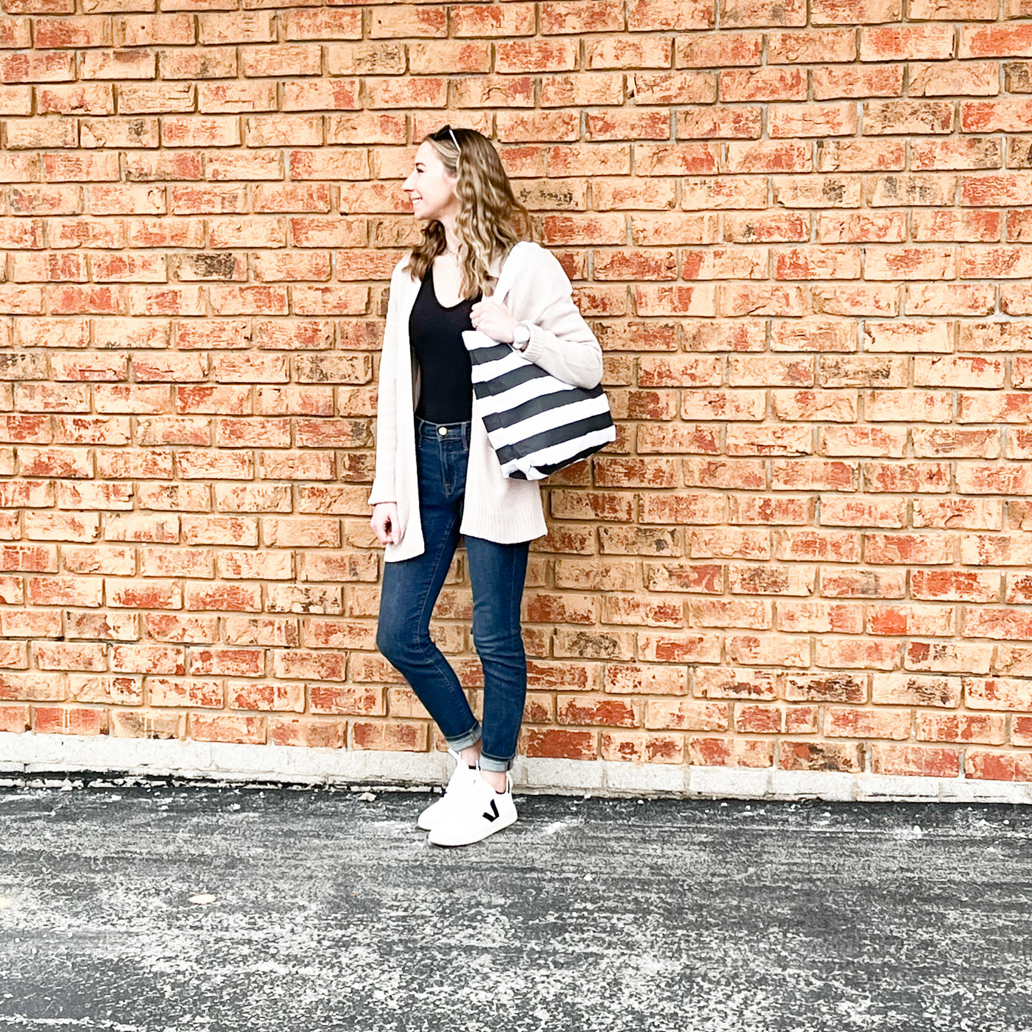 A smiling young woman standing on a blacktop parking lot. She is dressed in a black top, beige cardigan, blue jeans and white sneakers. She has sunglasses on top of her head, and a black and white bold striped tote bag on her shoulder. There is a brick wall in the background.