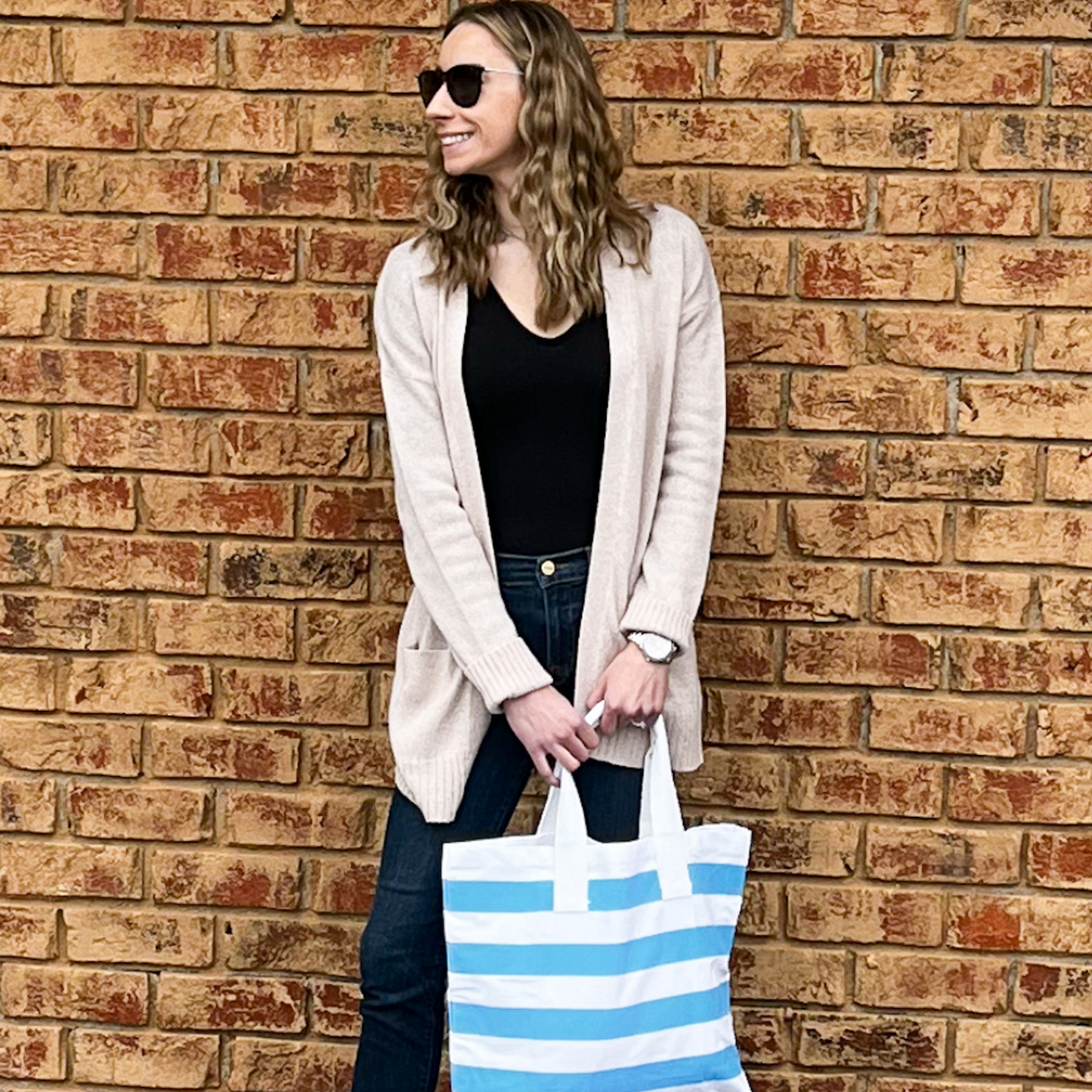 A smiling young woman is standing with her back to a brick wall. She is wearing a black top, beige cardigan, and blue jeans. She is wearing sunglasses and is using both hands to hold a blue and white bold striped tote in front of her.