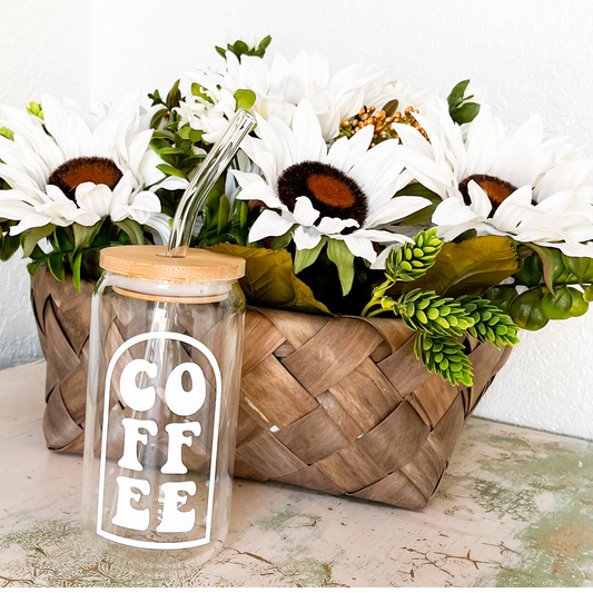 A glass can with a white vinyl design that is a thin outline with a curve on top. Inside the outline is the word coffee listed in two letter groups on top of each other. The glass can has a bamboo lid and a glass straw. in the background is a brown woven basket that holds white daisys and greenery.