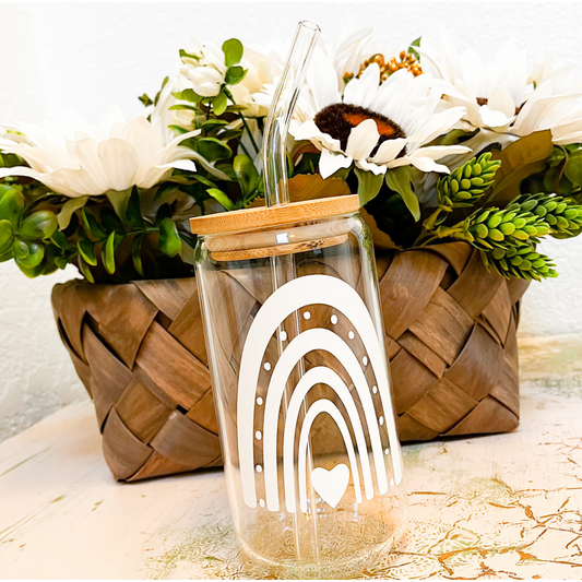 A glass can with a white vinyl rainbow design. The glass can has a bamboo lid and glass straw. In the background is a brown woven basket that is holding white daisys and greenery.