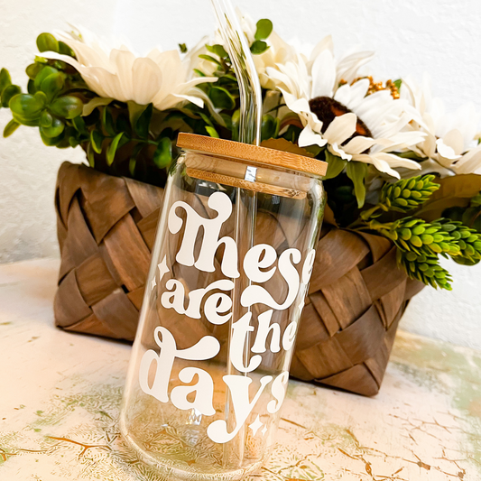 A glass can with a white vinyl decal that says "These are the days". The glass can has a bamboo lid and a glass straw. In the background is brown woven basket with a white daisy and greenery floral arrangement.